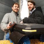 Two men stand outside two grain elevators with a Grain Weevil bin safety and management robot between them.