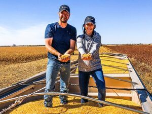 Levi and Jena Oschner of Double O Farms stand in a grain cart, holding out handfuls of corn and smile at the camera